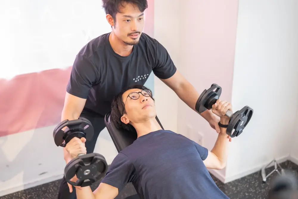 通い放題のパーソナルジムKARADA BESTAでパーソナルトレーニングを受ける30代男性。両手にダンベルを持ち、上を向いてあげようとしている。